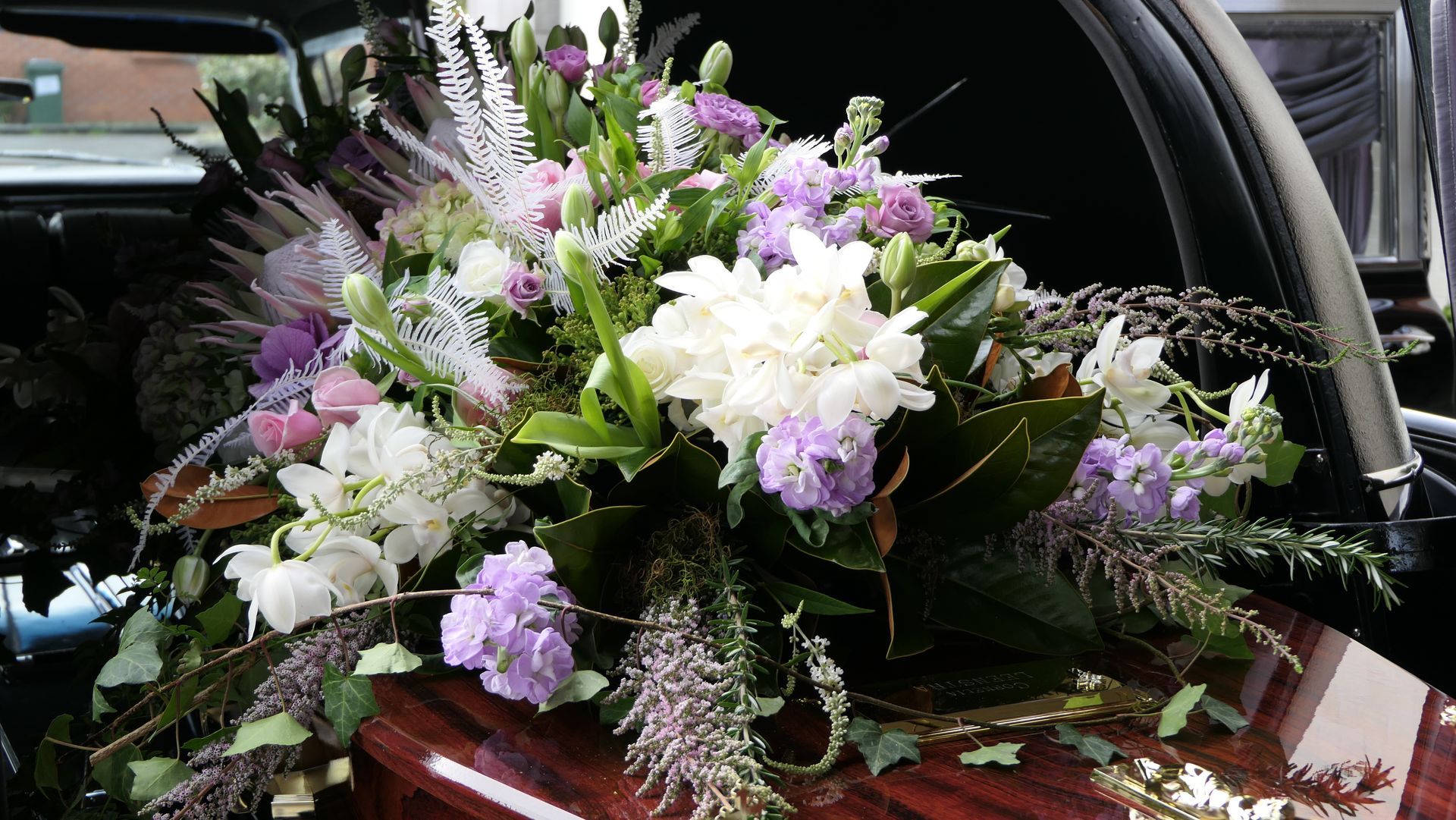 Fiori funebri bianchi, viola e verdi sopra una bara di legno scuro all'interno di un carro funebre.
