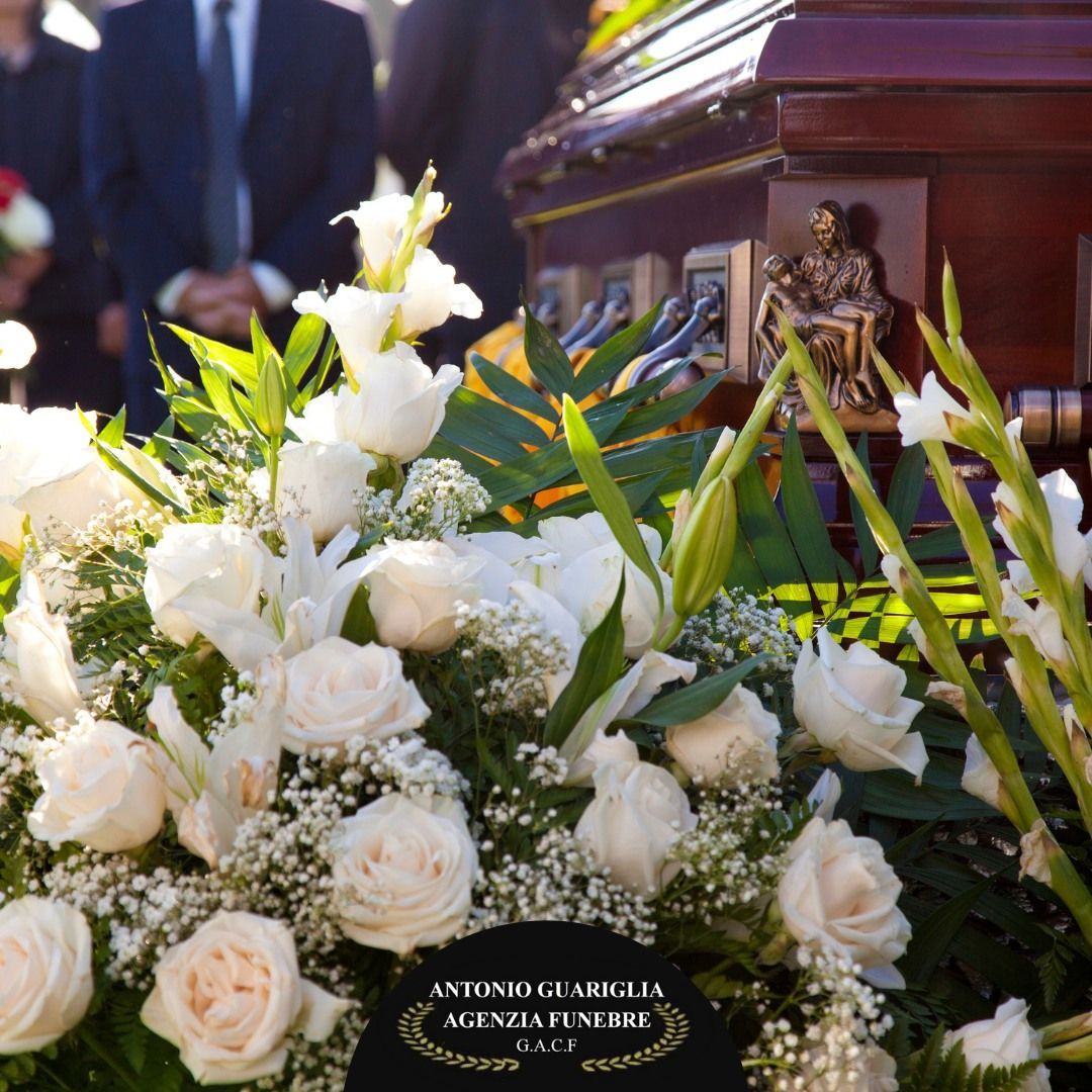 Fiori bianchi davanti a una bara durante un funerale, con persone in giacca e cravatta sullo sfondo.