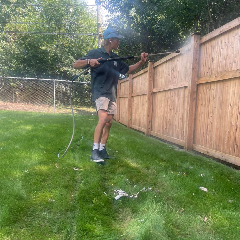 Person power washing a wooden fence in a grassy yard on a sunny day.