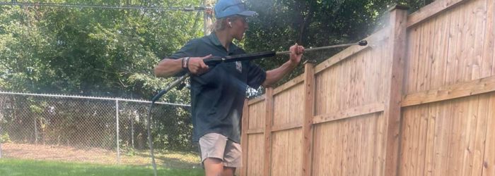 A person power washing a wooden fence outdoors in a sunny setting.