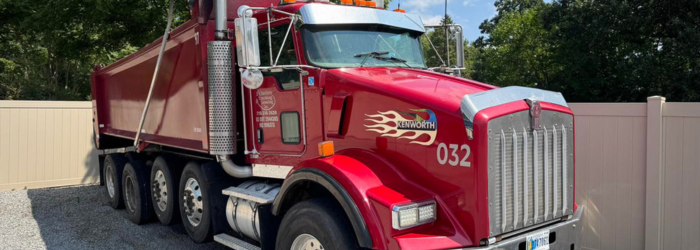 Red dump truck parked on a paved area next to a white fence. The truck has