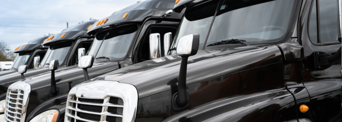 Row of black semi-trucks parked outdoors under a cloudy sky.