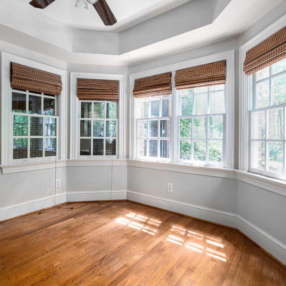 Empty room with hardwood floors, bay windows with woven shades, and a ceiling fan.