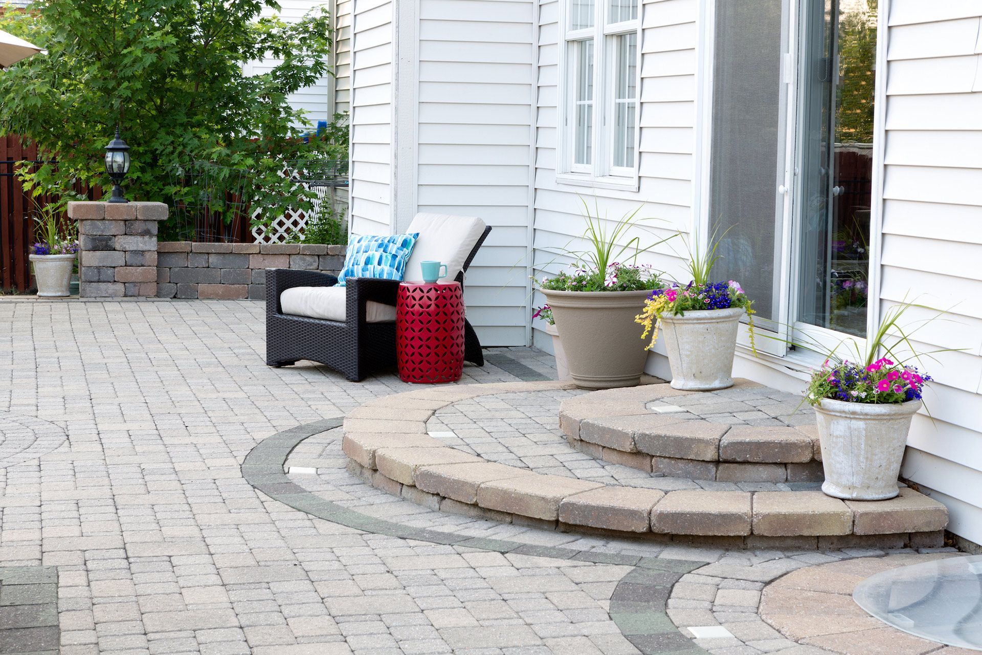 A landscape construction patio with a chair , table and potted plants in front of a white house.