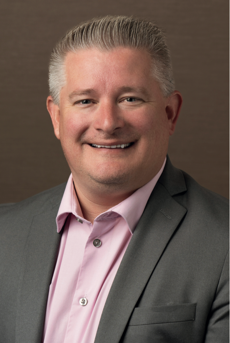 Man in gray blazer and pink shirt smiles, headshot against a brown background.