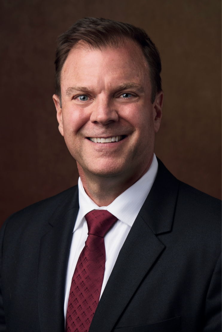 Man in a suit smiles, dark tie, against a brown background.