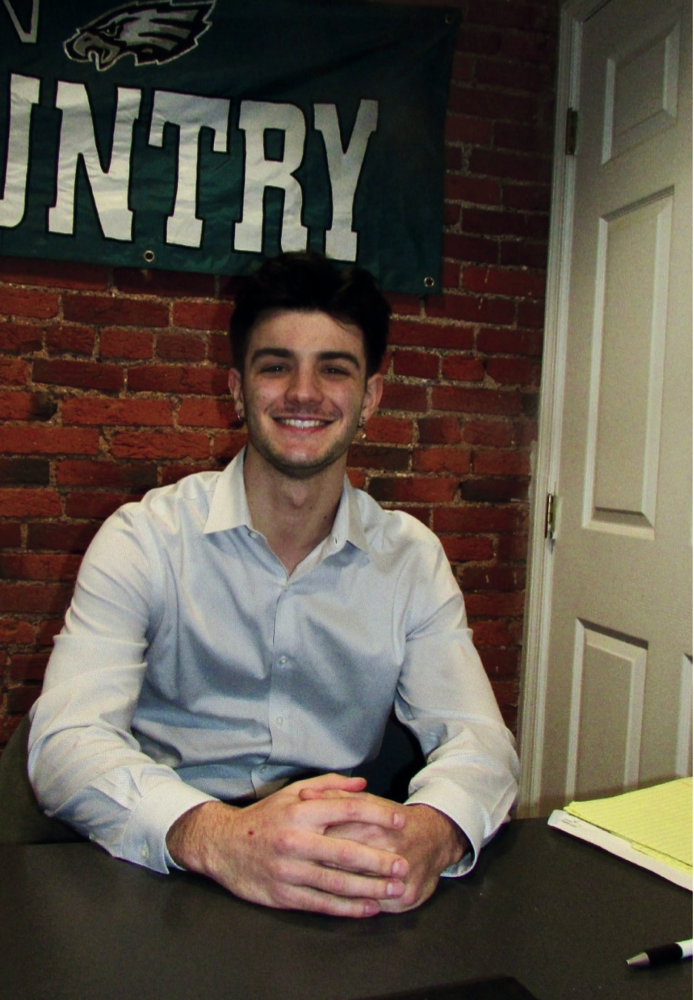 Man smiling, sitting at a desk, with hands clasped. Brick wall and white door in background. Eagles flag.