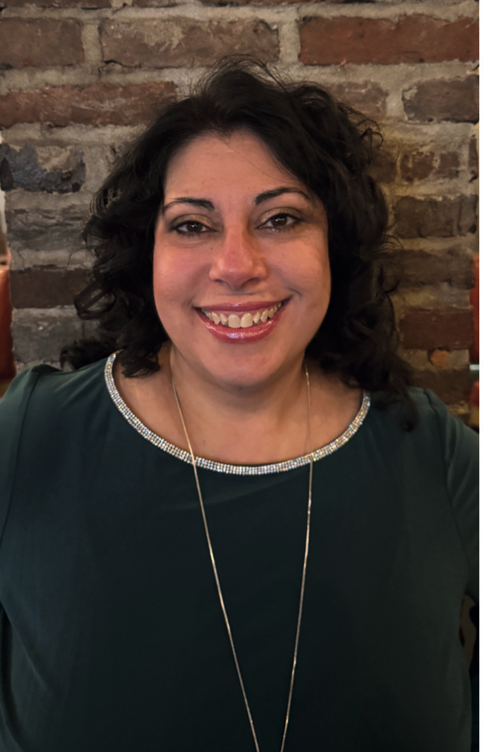 Woman with dark hair smiles, wearing a green shirt, necklace, in front of a brick wall.