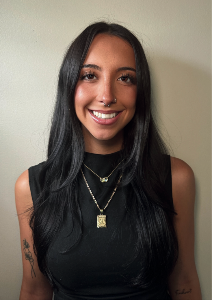 Woman with long, dark hair smiles, wearing a black top and gold necklaces, against a beige wall.