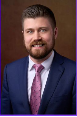 Man in blue suit, white shirt, and pink patterned tie, smiling against a brown backdrop.