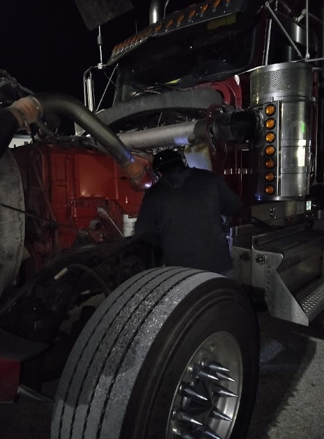 Mechanic working on a red semi-truck engine at night. One person leans in; tire in foreground.