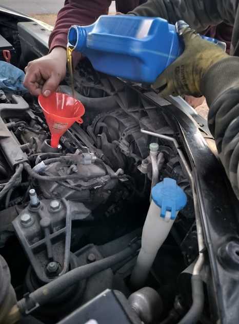 Hands pouring oil into a car engine with a funnel. Blue oil container, red funnel, and engine visible.