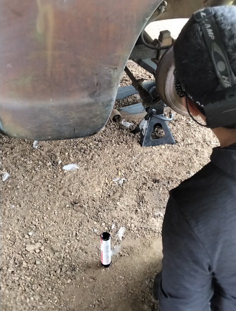 Person looking under a vehicle, supported by a jack stand, bottle on the ground.