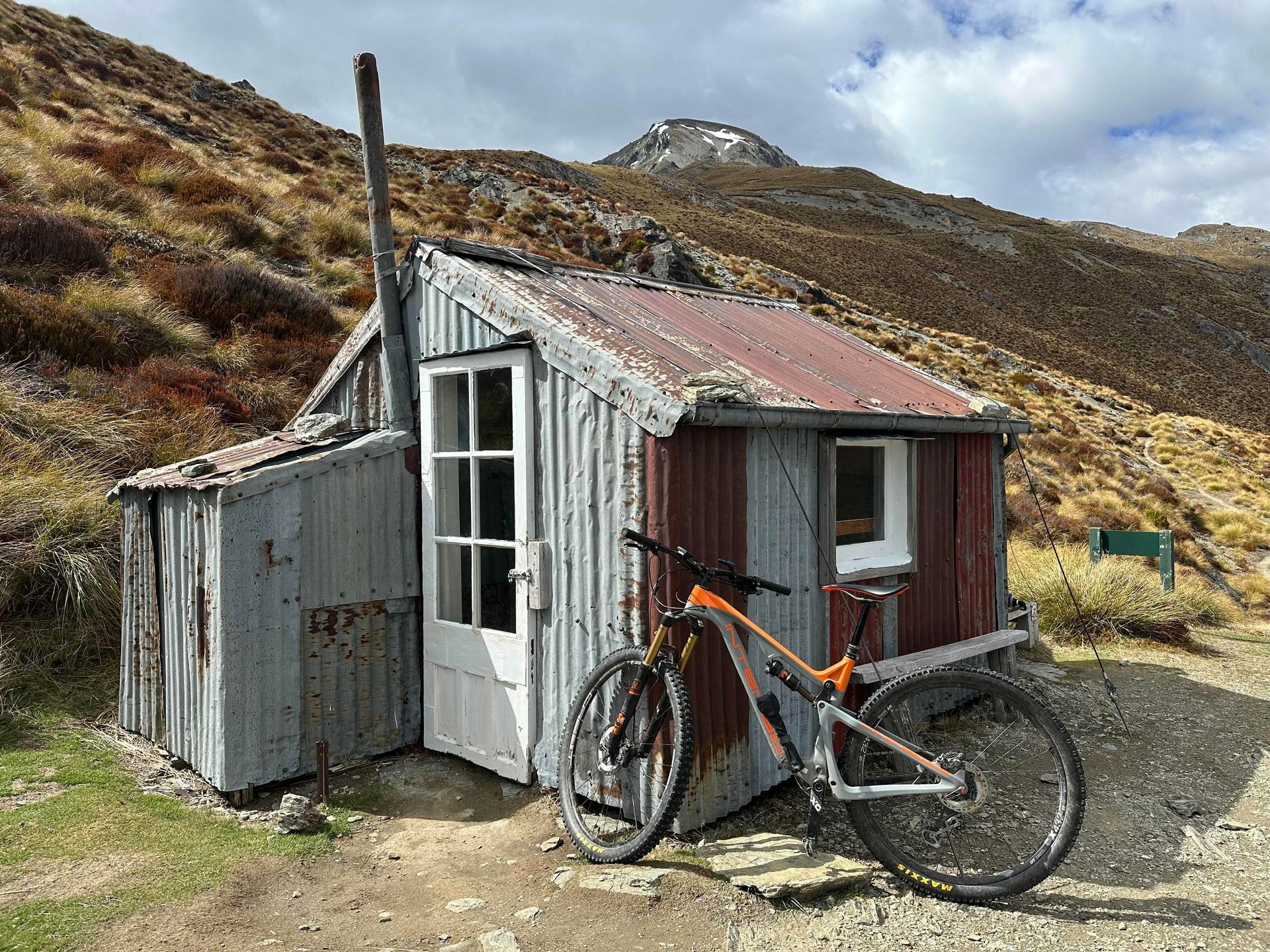 Mountain bike leans against a weathered, corrugated iron hut on a hillside.