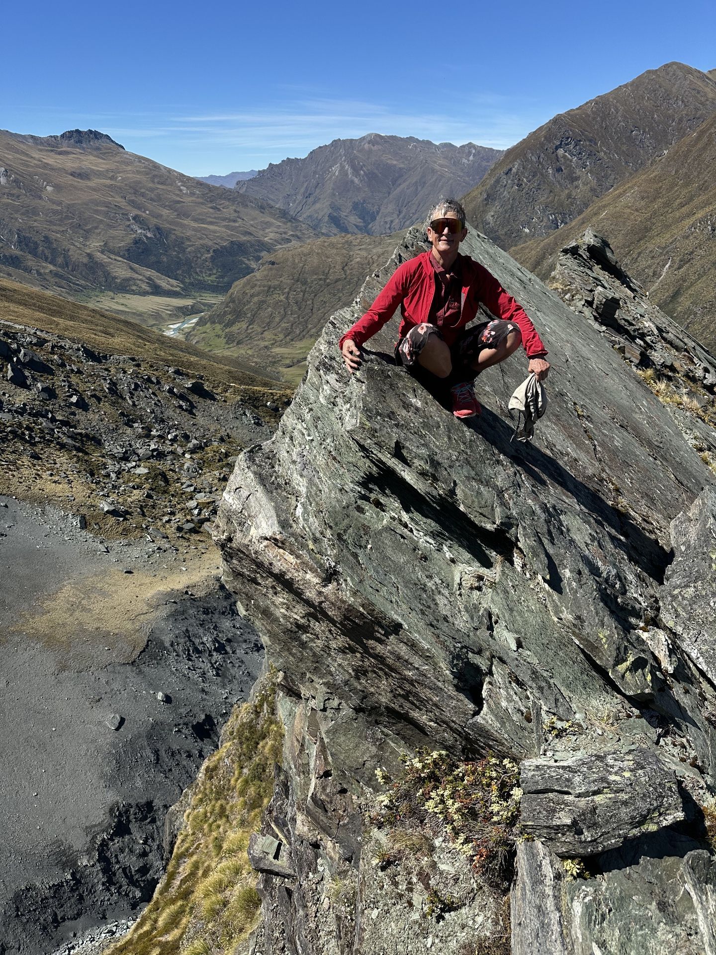 Person crouching on rocky ridge overlooking valley, wearing red shirt, blue sky.