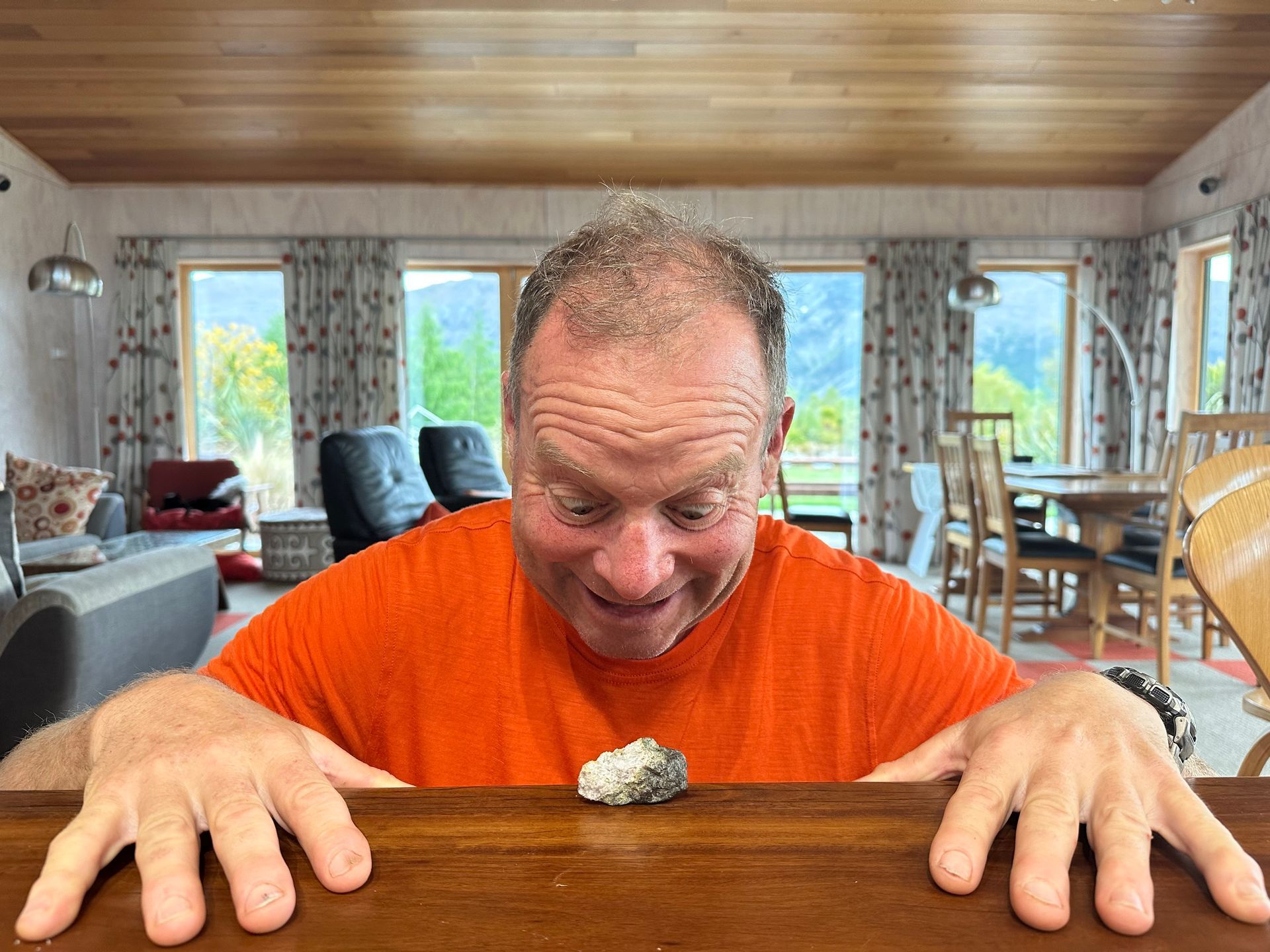 Man in orange shirt examines small rock on table in a bright, modern room.