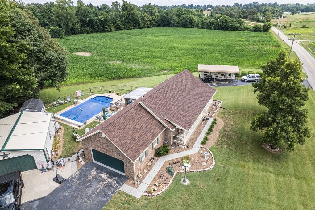 Aerial view of a house with a pool, surrounded by greenery and farmland, with a paved driveway.