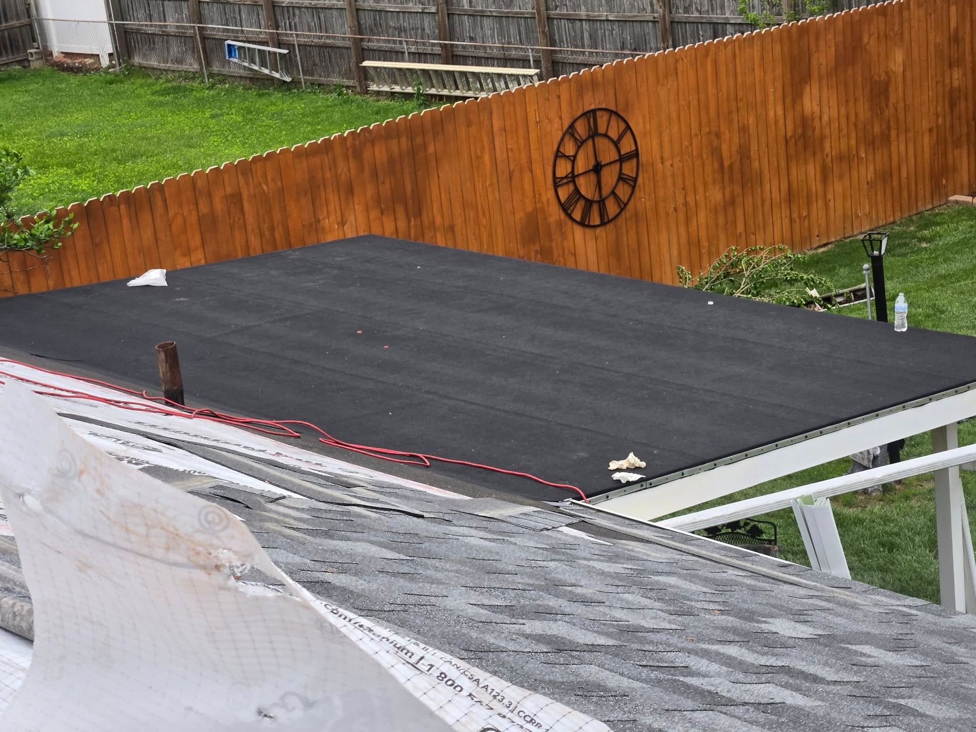 Flat roof with black surface, adjacent to a wooden fence with a clock. Grass and a white support structure are also visible.
