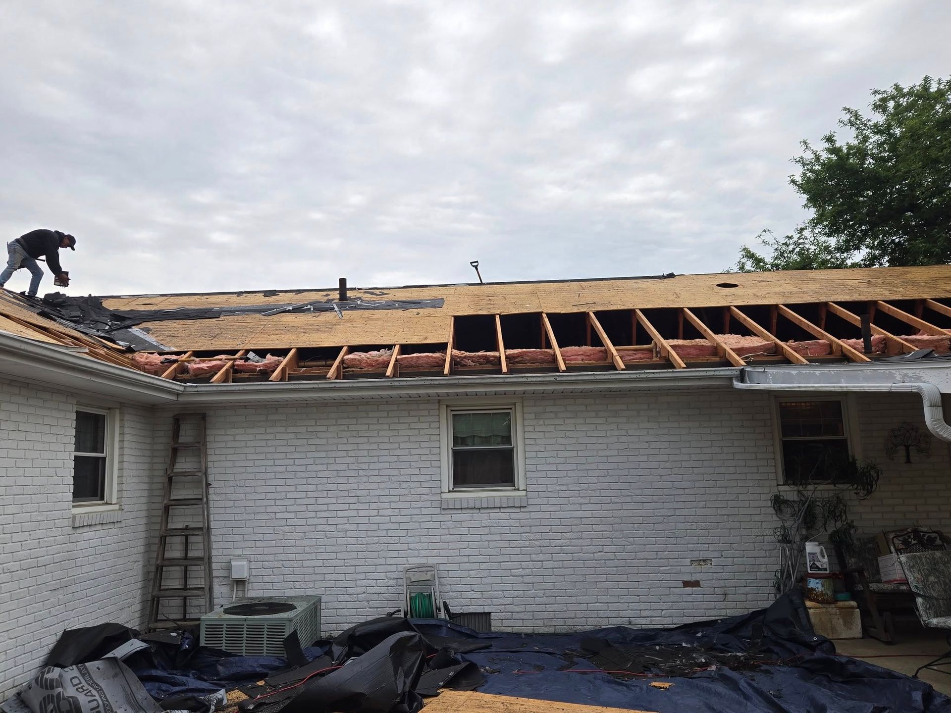 A worker on a roof removing shingles and repairing wooden framing; exterior of a brick house.