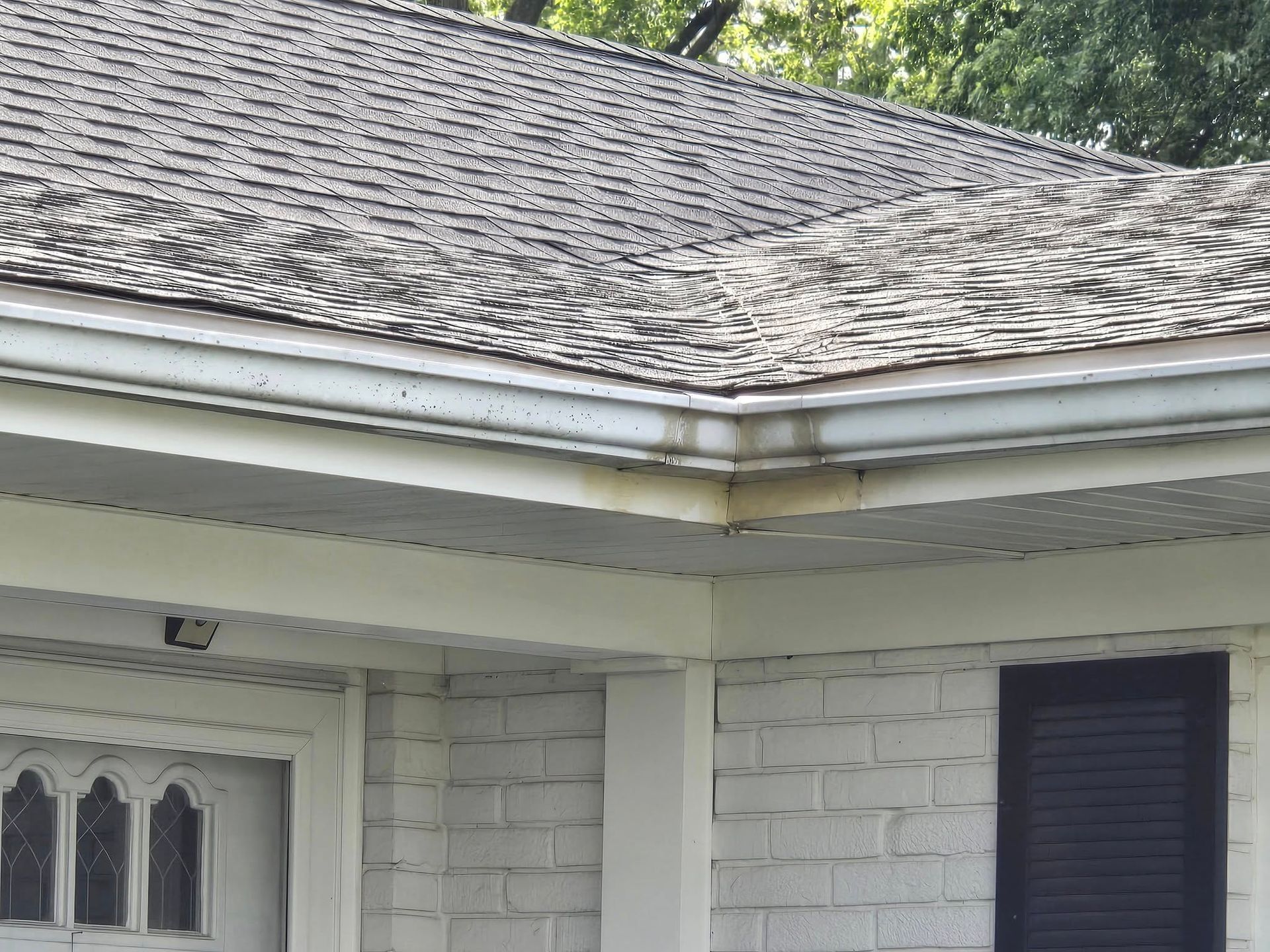 Corner of a house with a gray roof and white gutters, near a dark shuttered window.