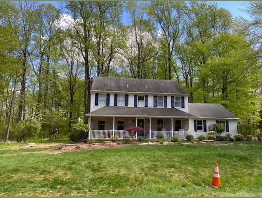 A two-story white house with blue shutters and a front porch, nestled among lush green trees under a bright sky.