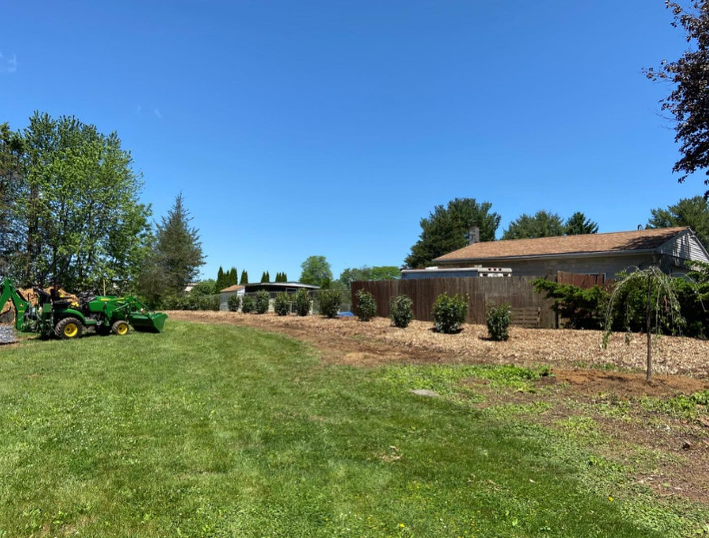 A green tractor sits on a sunny, mowed lawn next to a landscaped garden bed with mulch, small shrubs, and a house.