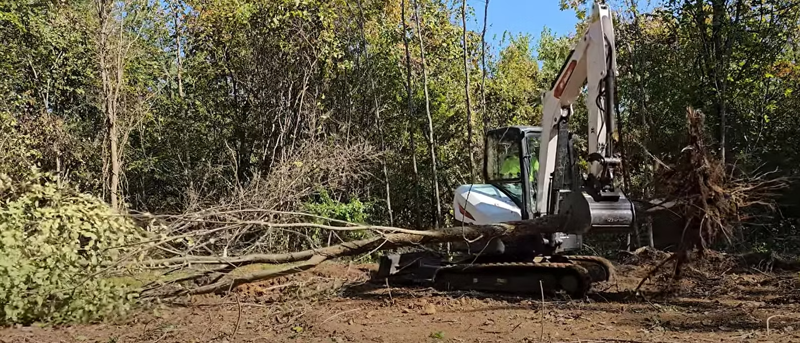 A white excavator clearing fallen tree branches and debris from a wooded area on a sunny day.