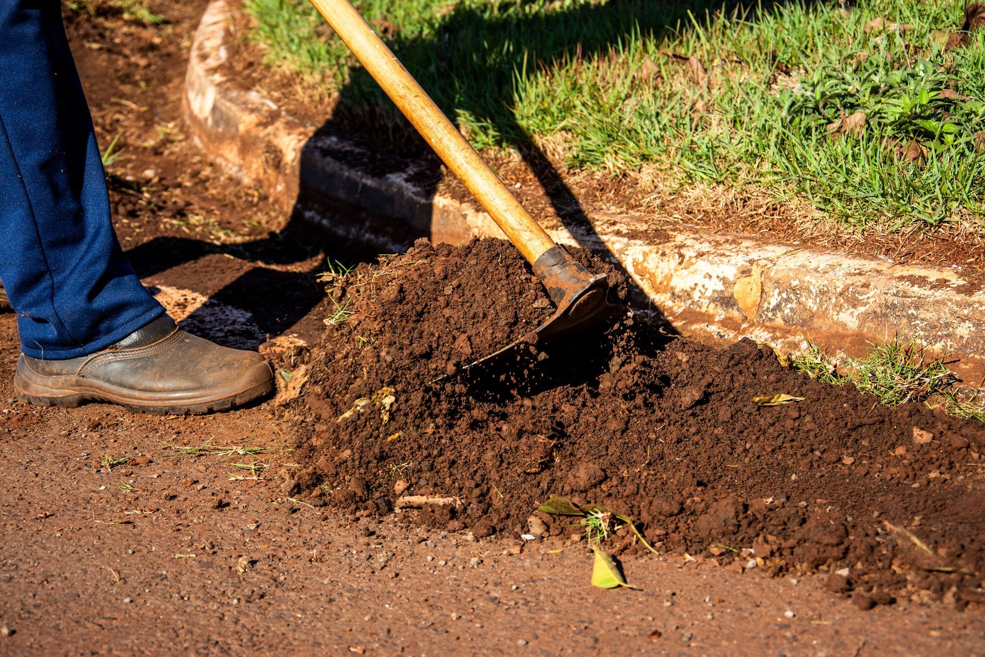 Person shoveling soil beside a curb, with dirt piled on the roadside and grass nearby