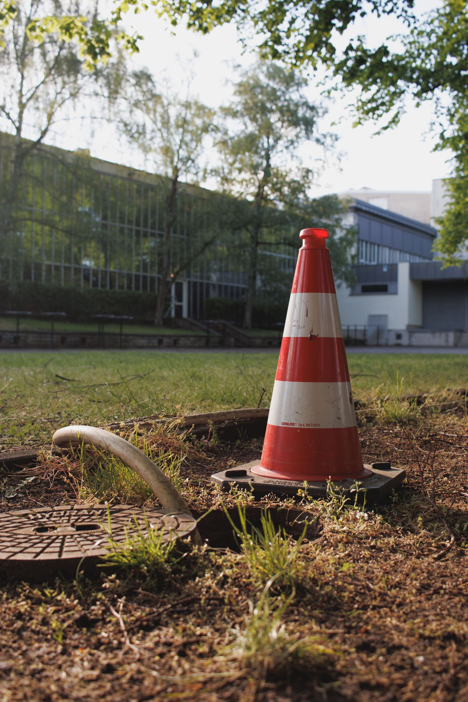 Orange-and-white traffic cone on grass near a building and trees, with a hose in the foreground