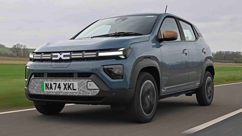Blue Dacia Spring EV driving on a road. License plate covered, with brown roof accents and black trim.