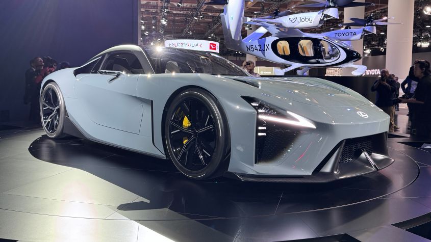 Gray sports car on display, with a flying vehicle in the background. Black wheels, yellow brake calipers, and modern design.