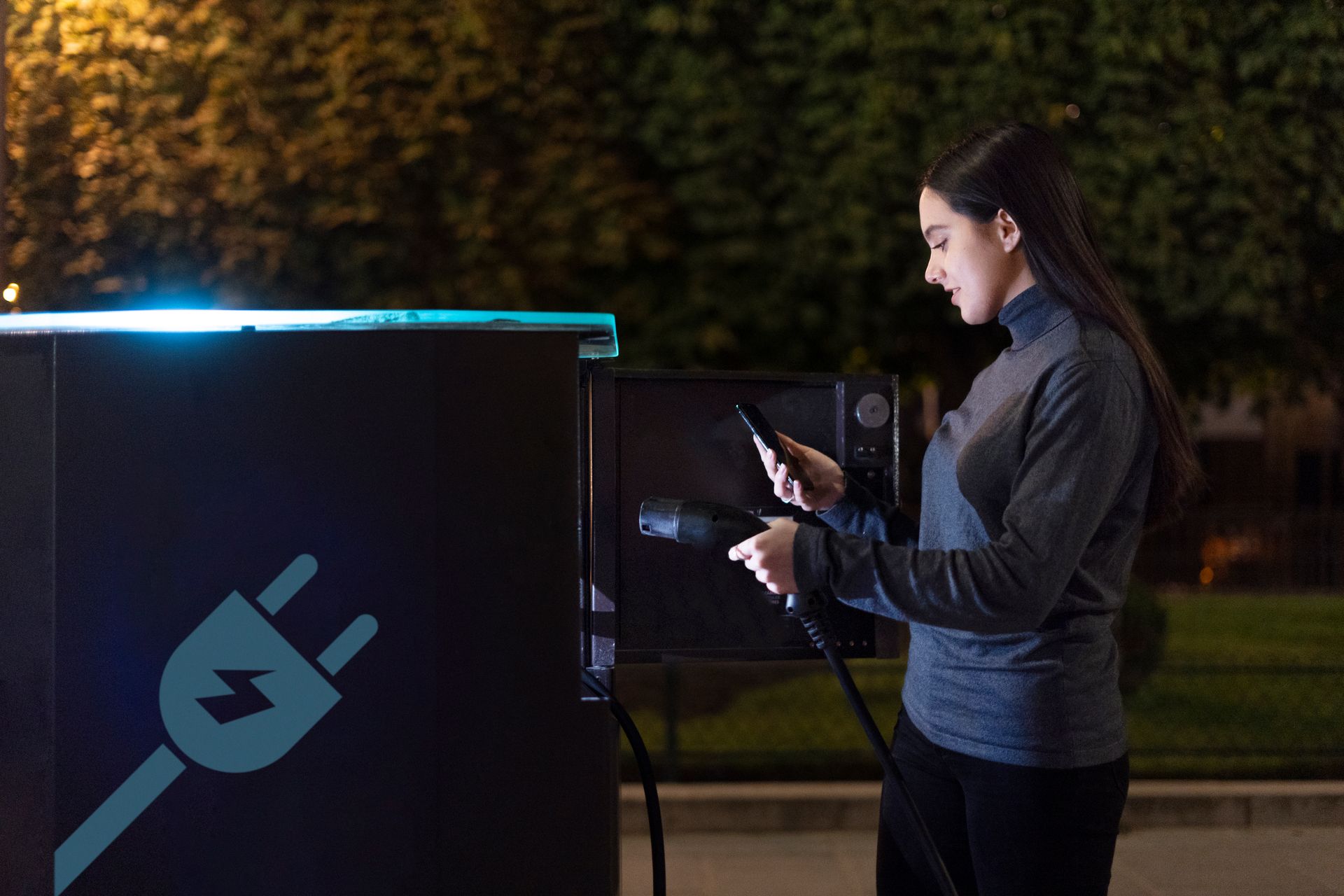 A woman is charging her electric car at a charging station while looking at her phone.