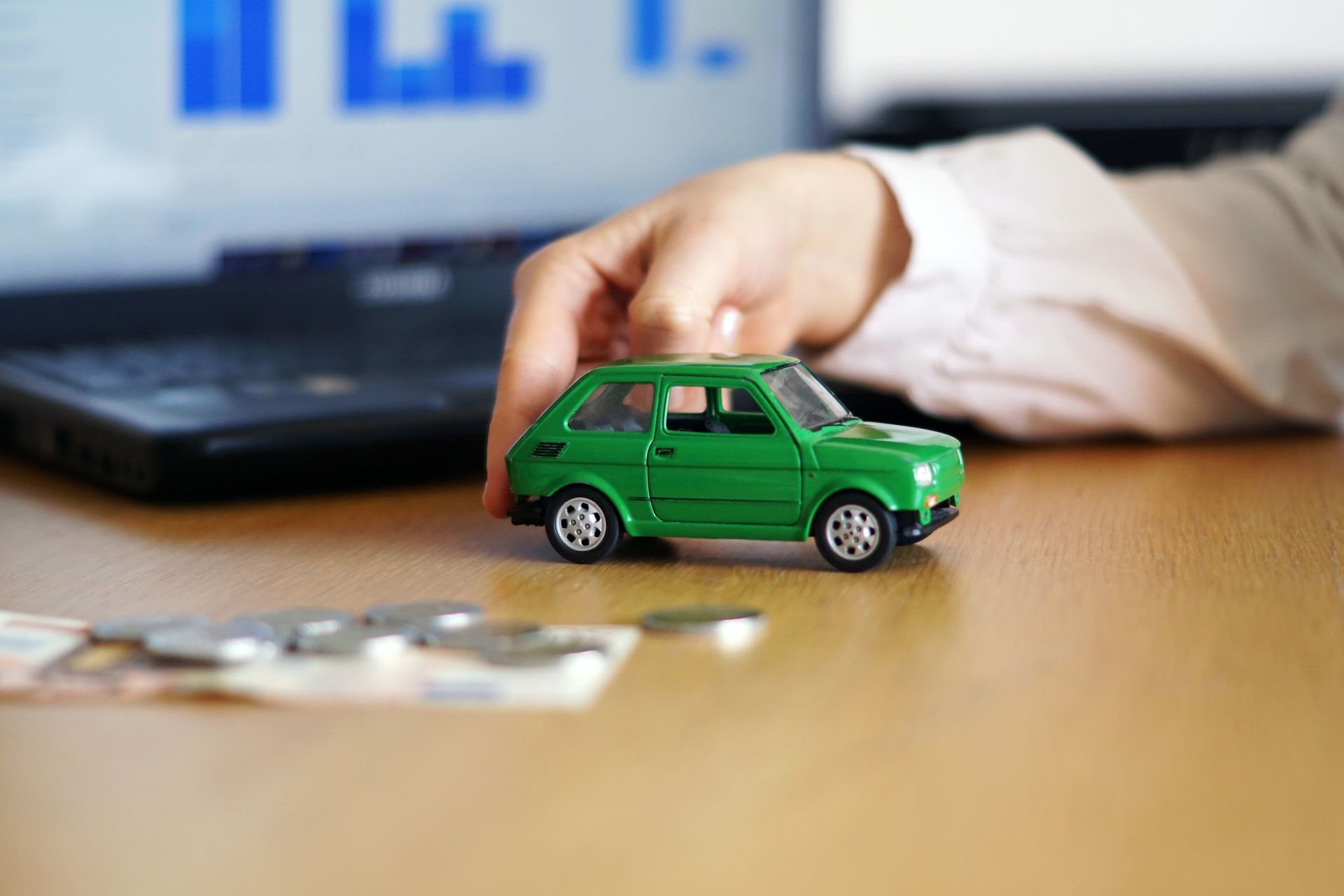Hand holding a green toy car on a desk with money and a laptop in the background.