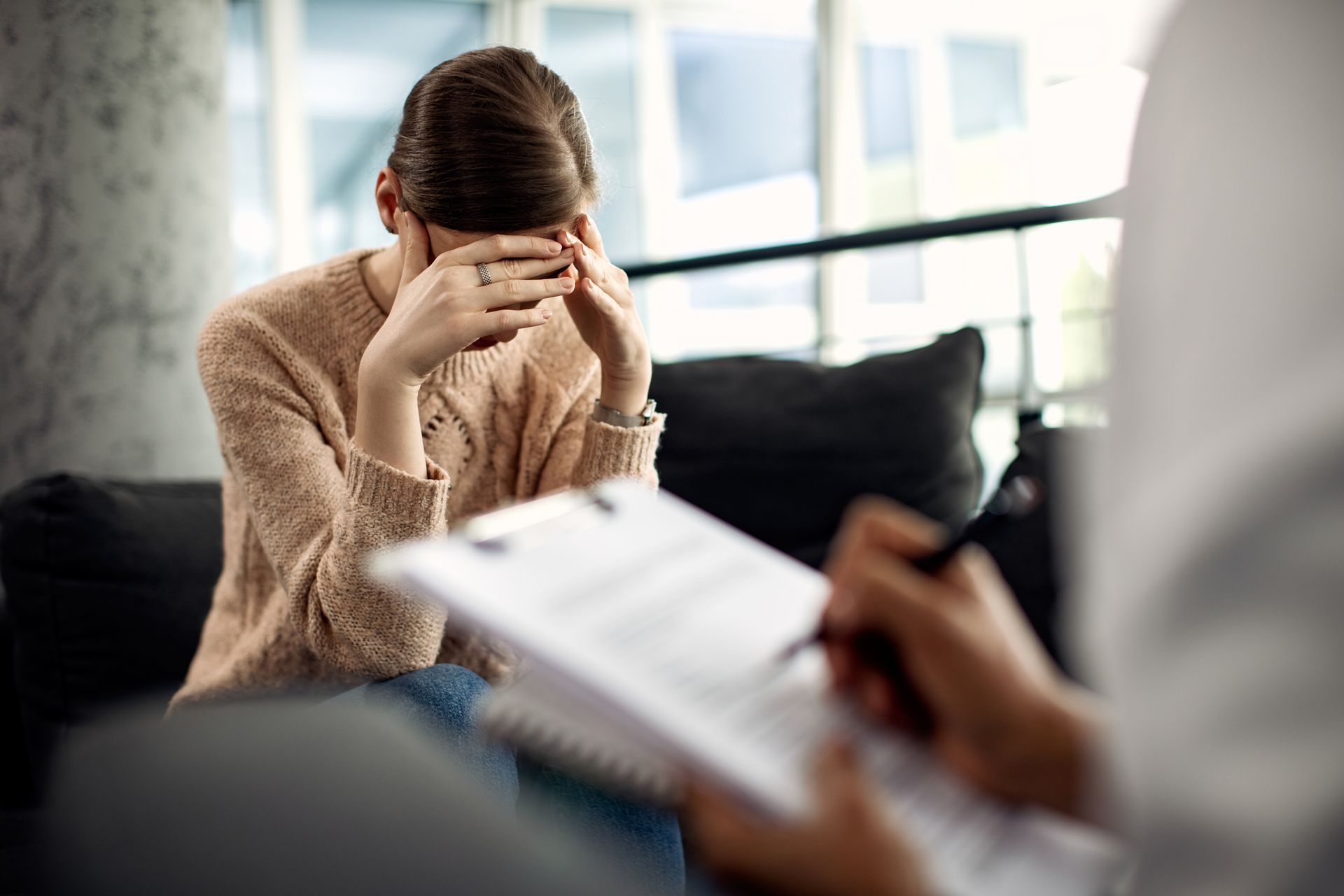 A woman is sitting on a couch with her head in her hands while a man writes on a clipboard.