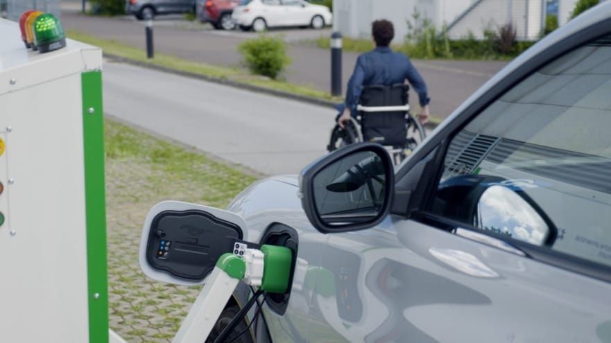 A man in a wheelchair is standing next to an electric car that is being charged.