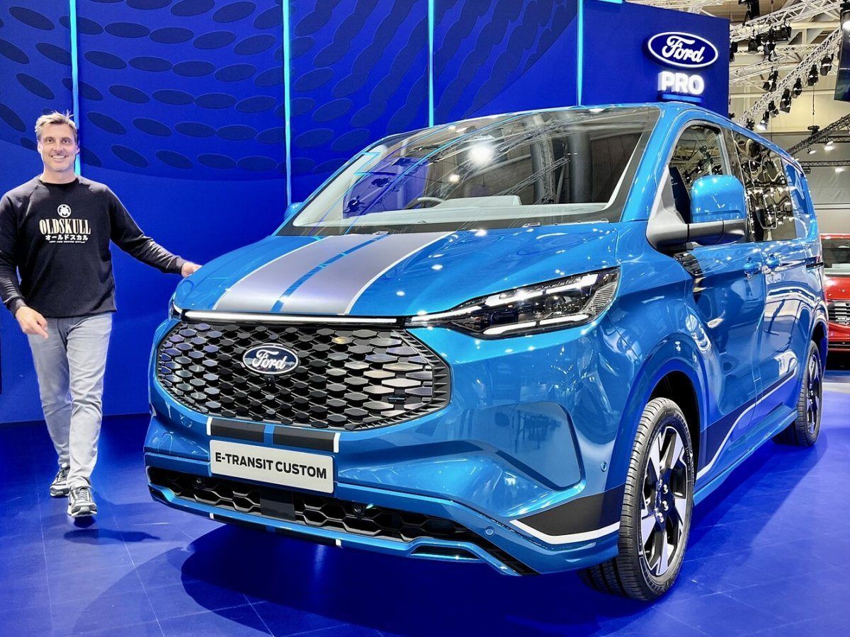 A man is standing next to a blue Ford e-Transit Custom at a car show. 