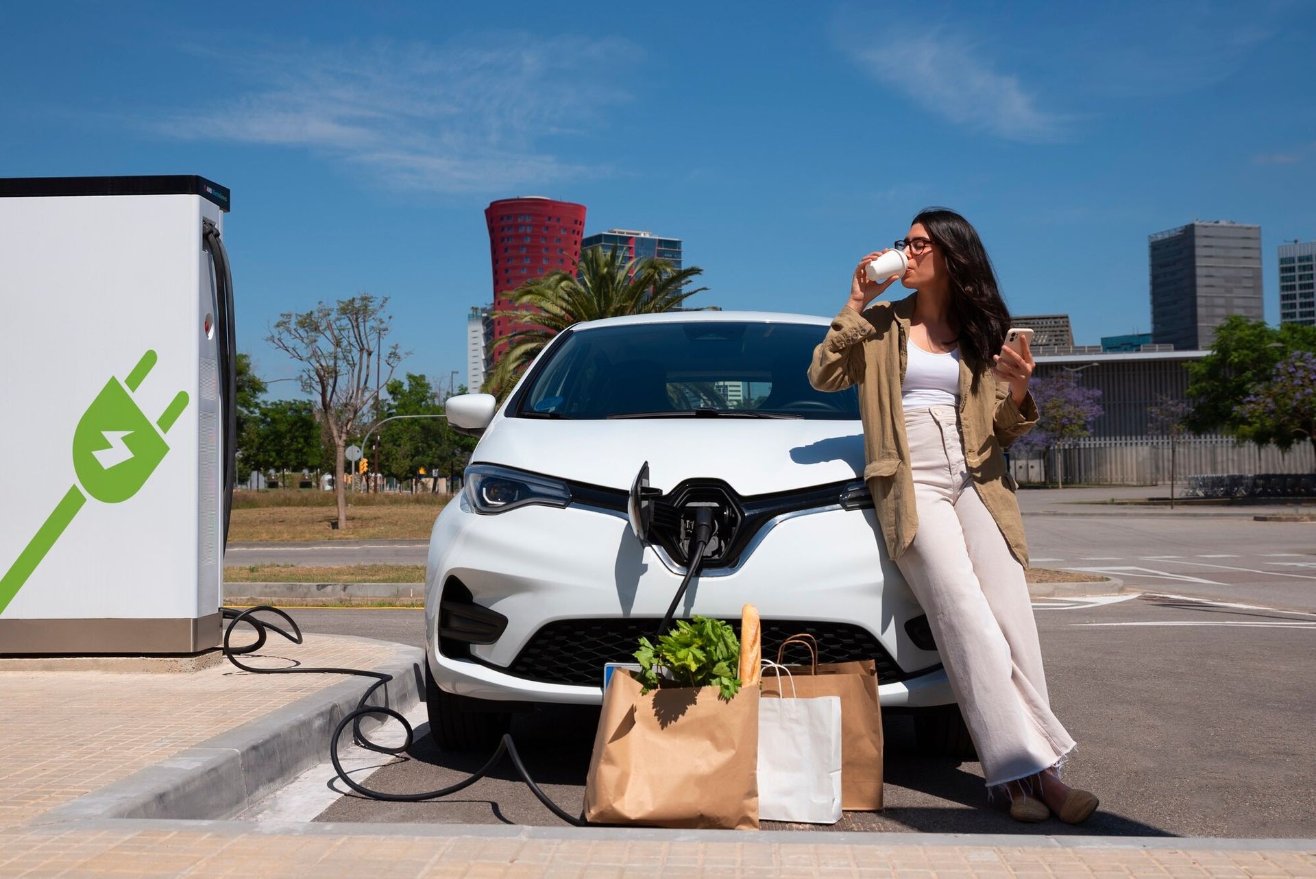 A woman is sitting in front of an electric car at a charging station.