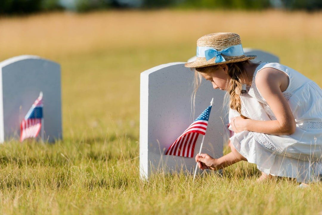 Woman places an American flag at a grave in a sunny cemetery.