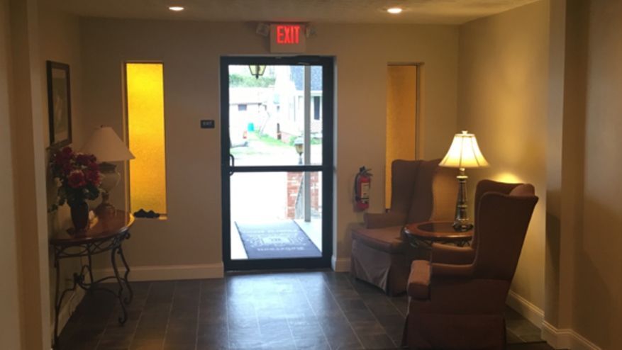 Hallway with exit door, two chairs, and table with flowers and lamps. Yellow panels on the walls.
