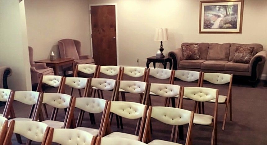 Rows of folding chairs face a room with a sofa, end tables, and a framed picture; a funeral home.