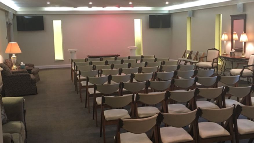 Rows of chairs face a stage in a beige-walled room. Two TVs hang on the wall.