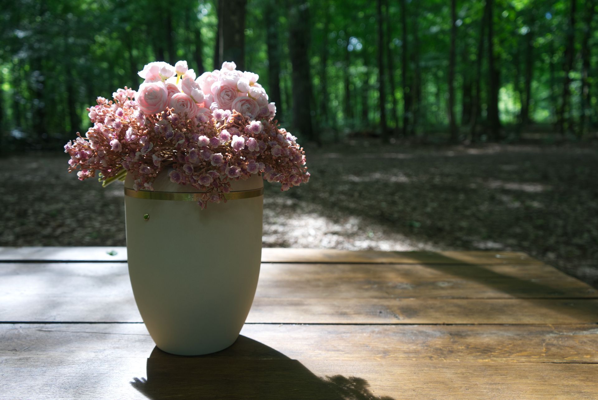 Vase of pink flowers on a wooden table in a sunlit forest clearing.