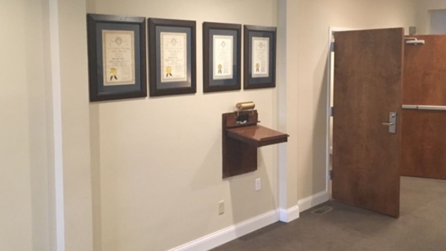 Hallway with framed documents on wall above a small desk, next to a door.
