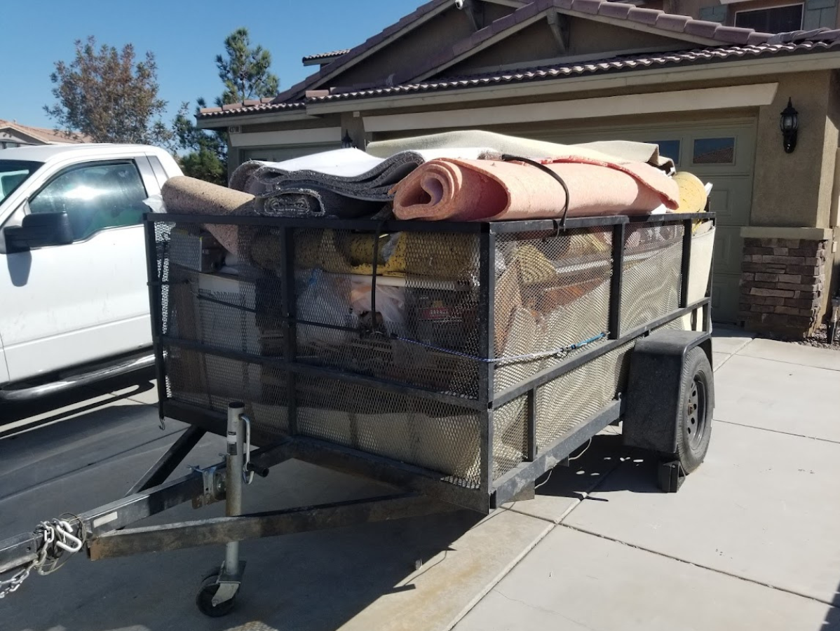 A trailer loaded with rolled-up rugs, wood, and other items, parked in front of a house next to a truck.