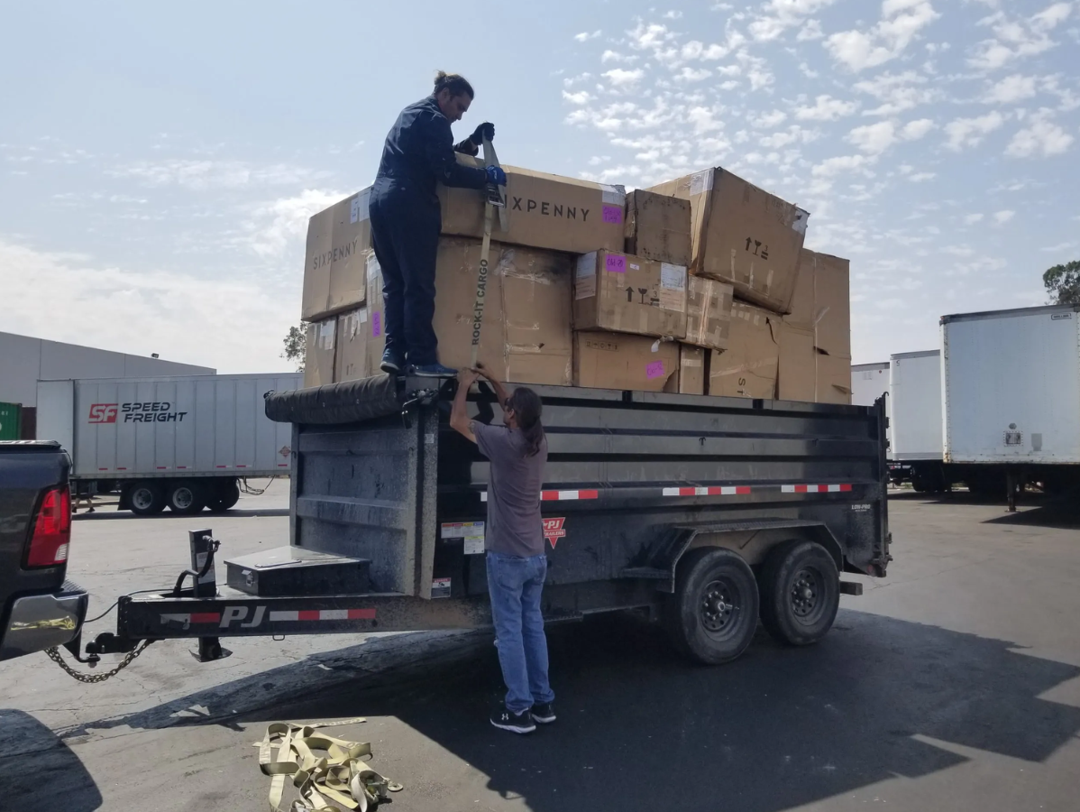 Two people loading cardboard boxes into a dump trailer in an outdoor parking lot.