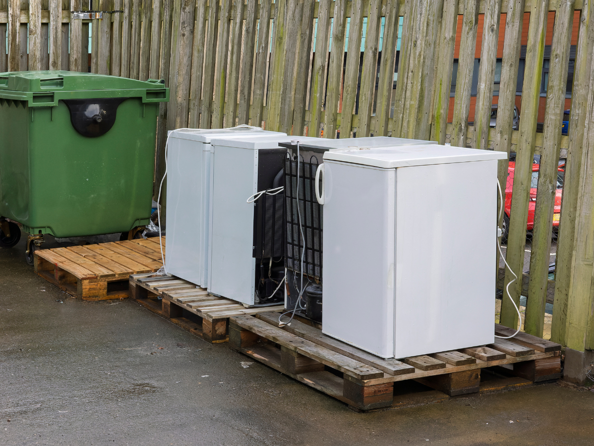 Green trash bin and white refrigerators on pallets next to wooden fence.