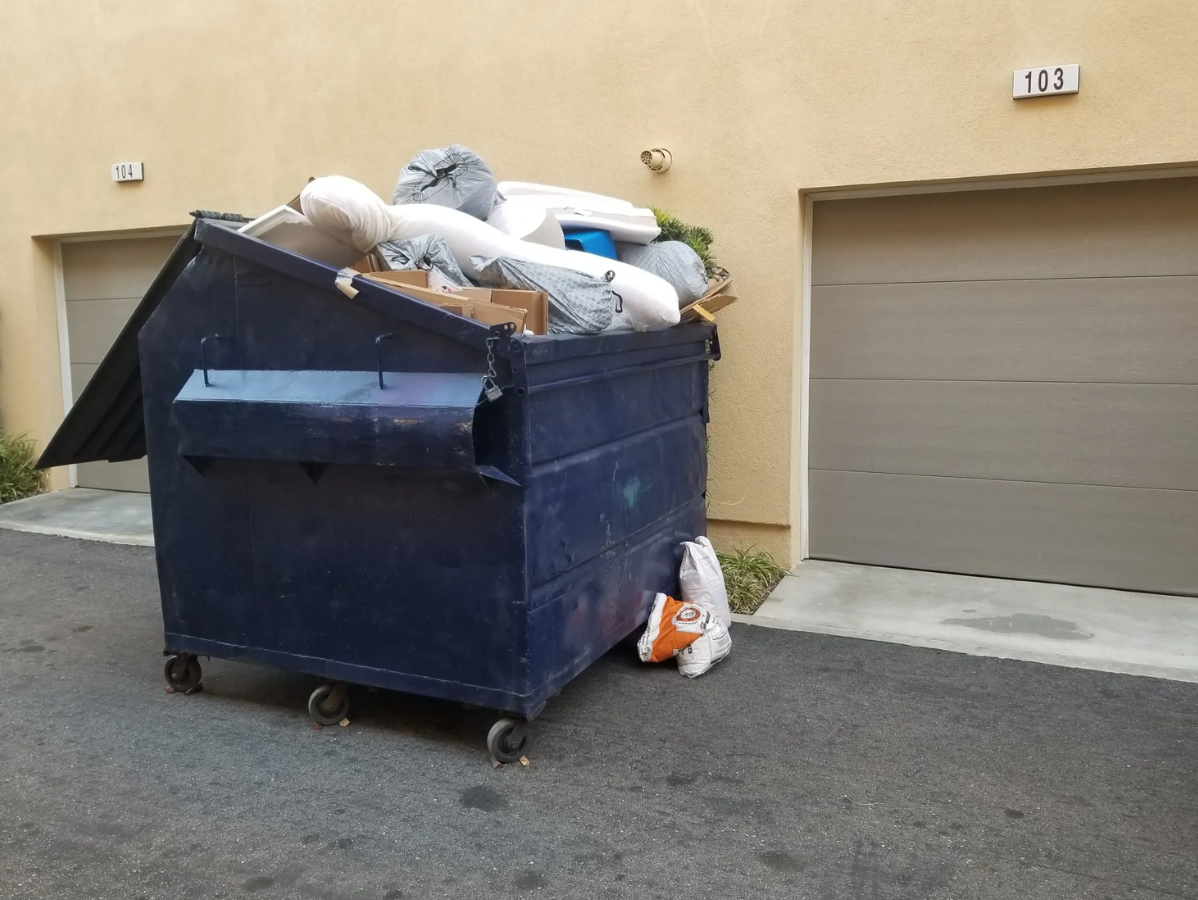 Overflowing blue dumpster next to beige garage doors and tan building exterior.