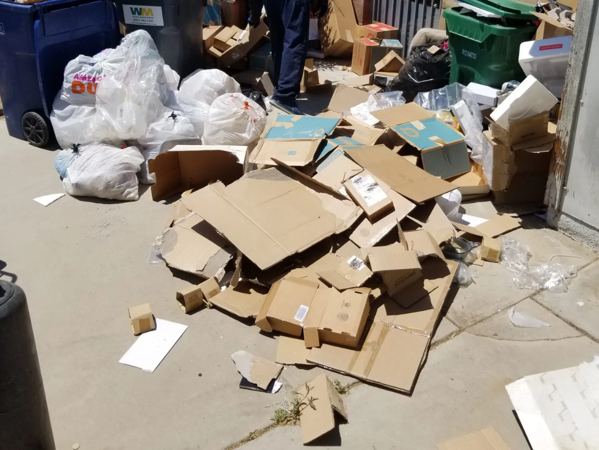Pile of flattened cardboard boxes and trash bags on a sidewalk, next to trash bins.