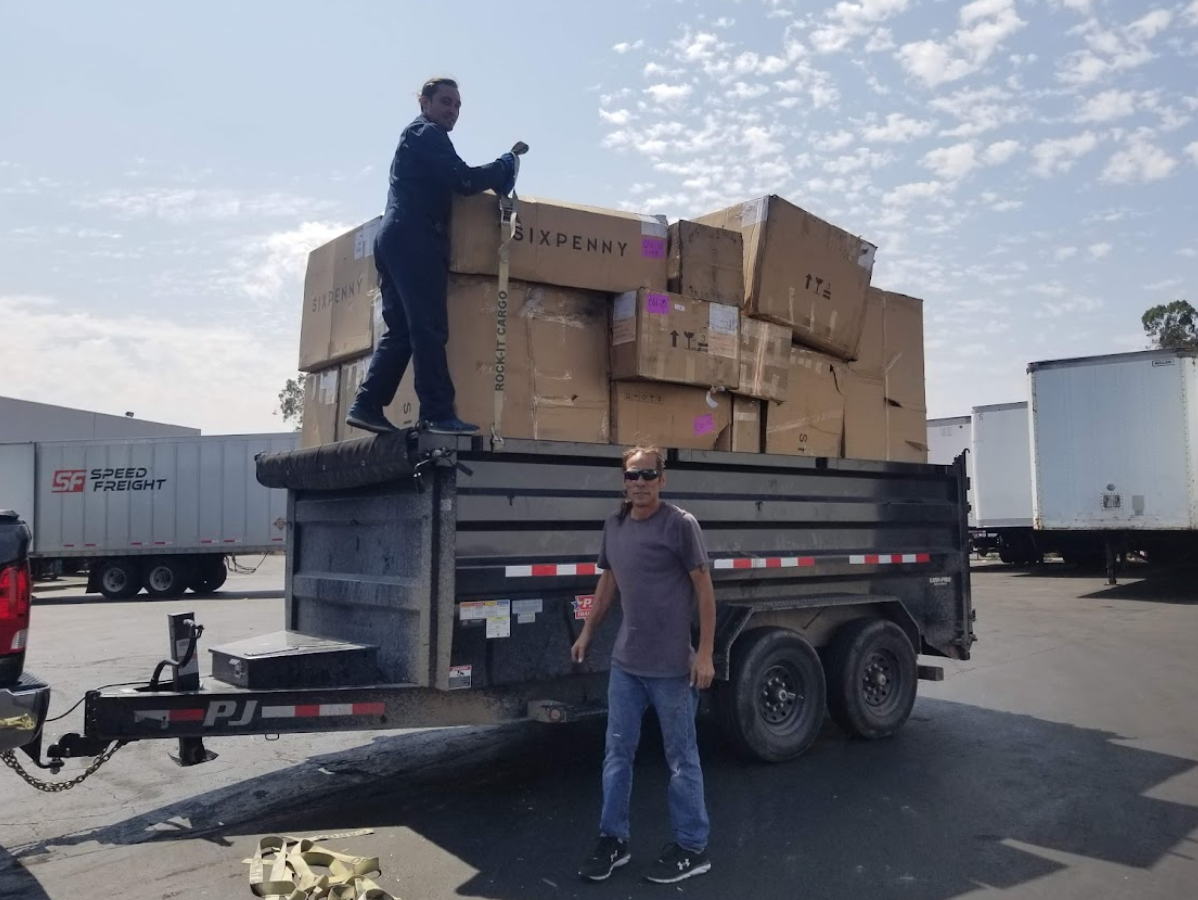Two people loading boxes onto a trailer in a parking lot. Boxes stacked high, blue sky background.