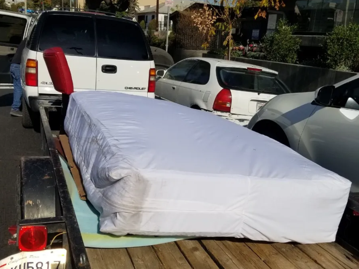 A mattress wrapped in white plastic is on a trailer, attached to a white SUV.